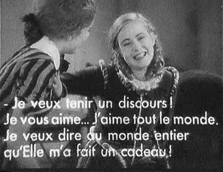 Scène de film en noir et blanc montrant deux femmes, dont l&rsquo;une sourit largement. Le sous-titre indique : « - Je veux tenir un discours ! Je vous aime&mldr; J&rsquo;aime tout le monde. Je veux dire au monde entier qu&rsquo;Elle m&rsquo;a fait un cadeau ! »