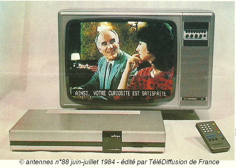 Un ancien téléviseur cathodique associé à un décodeur Thomson et une télécommande. L&rsquo;écran montre un homme et une femme avec le sous-titre en majuscules : « Ainsi, votre curiosité est satisfaite. ».