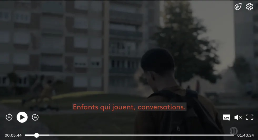 Capture d&rsquo;écran d&rsquo;un lecteur vidéo montrant une personne de dos avec un sac à dos, marchant dans un quartier résidentiel avec des immeubles en arrière-plan. Le sous-titre descriptif indique en rouge : « Enfants qui jouent, conversations. ».